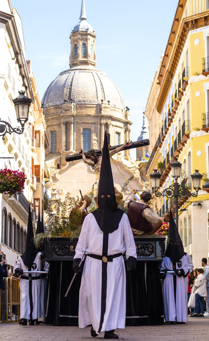 A religious Easter procession in Zaragoza with hooded participants and a historic dome background.